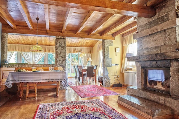 A rustic interior with a stone fireplace, wooden beams, a dining area with a table and chairs, and a view through a window.