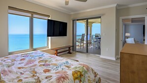 Een slaapkamer met een bed, een plafondventilator, een televisie, een bankje, een balkon met een tafel en stoelen, en uitzicht op de oceaan.