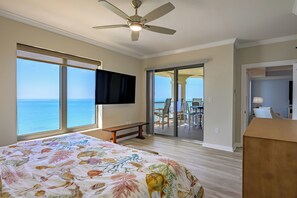 Ein Schlafzimmer mit Bett, Deckenventilator, Fernseher, einer Bank, einem Balkon mit Tisch und Stühlen und Meerblick.