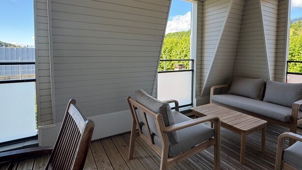 A deck with a wooden chair, a small table, and a sofa, overlooking a body of water and greenery. - Bright apartment with mountain view - 4 guests (Aix-les-Bains)