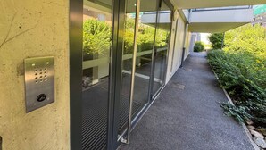 A corridor with glass doors, a metal panel with a keypad, and a pathway leading to a building entrance. - Bright apartment with mountain view - 4 guests (Aix-les-Bains)