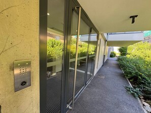 Een gang met glazen deuren, een metalen paneel met een keypad, en een pad naar de ingang van een gebouw.