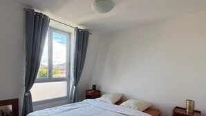 A bedroom with a bed, two bedside tables, a window with curtains, and a view of buildings outside. - Bright apartment with mountain view - 4 guests (Aix-les-Bains)