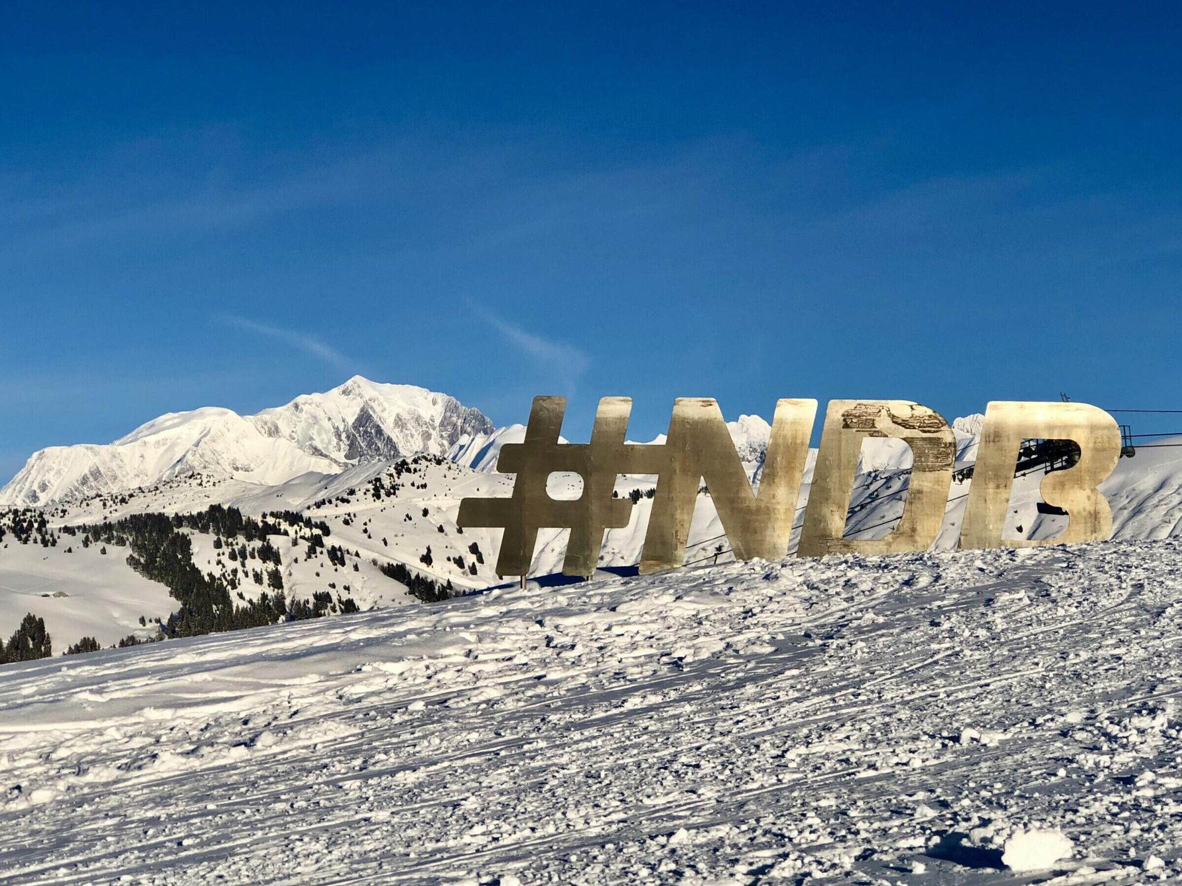 Un paysage montagneux enneigé, avec un grand panneau portant le hashtag « #NDR » au premier plan.