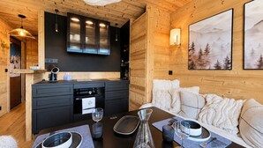 L’intérieur chaleureux d’un chalet avec un plafond en bois, un coin cuisine équipé d’appareils modernes, une table à manger pour deux personnes et un canapé agrémenté de coussins décoratifs.