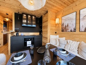 Ein gemütliches Blockhausschlafzimmer mit Holzbalkendecke, einer Küchenzeile mit modernen Geräten, einem Esstisch für zwei Personen und einer Couch mit dekorativen Kissen.