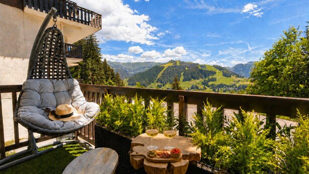 Un balcon agrémenté d’un fauteuil suspendu, une petite table avec de la nourriture et des boissons, et une vue sur des collines verdoyantes et des montagnes.