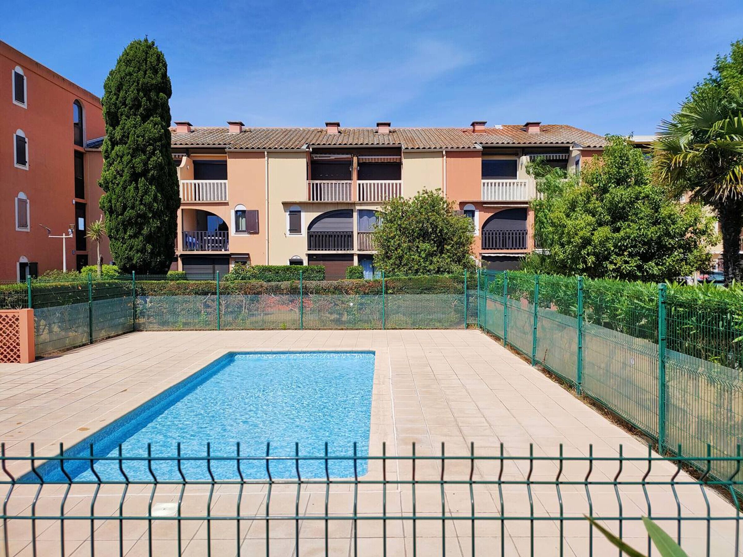 A swimming pool with a tiled deck, surrounded by a green fence, and multi-story buildings with balconies in the background.