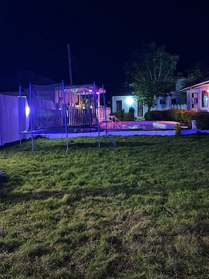 Een achtertuin met een trampoline, een klein speelhuisje en een boom.