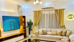 A living room with a beige sofa, a glass coffee table, a TV mounted on the wall, and a ceiling fan.