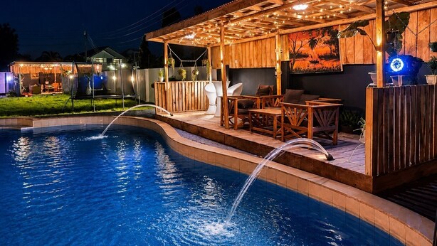 A pool area with a wooden deck, seating, and a covered patio with string lights.