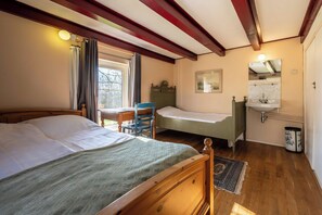 Un dormitorio con cama de madera, un escritorio, una silla, una mesita de noche pequeña, una ventana con cortinas, un espejo, un lavabo y un cubo de basura.