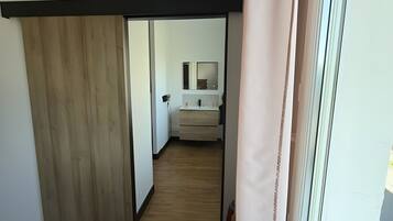Une pièce avec un plancher en bois, une chaise noire, une fenêtre avec un rideau et une salle de bain visible, équipée d’un miroir et d’un lavabo.