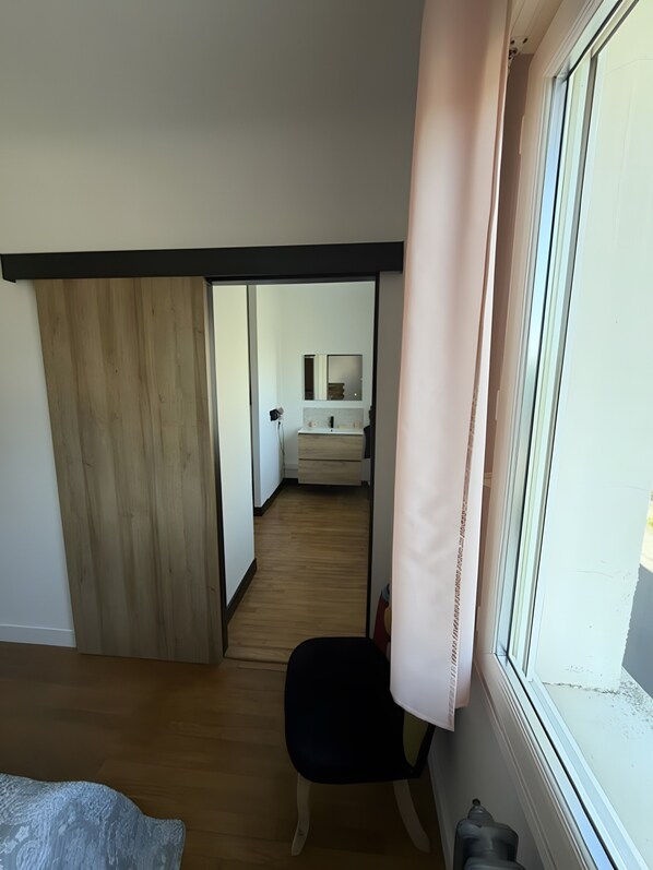 A room with a wooden floor, a black chair, a window with a curtain, and a visible bathroom with a mirror and sink.
