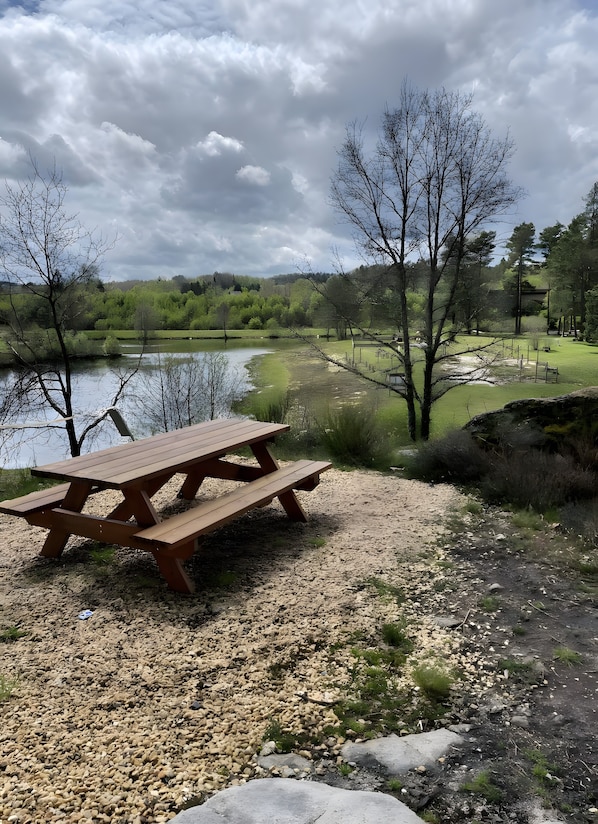 Una mesa de picnic junto a un lago, con un sendero de grava, árboles y una zona cubierta de césped.