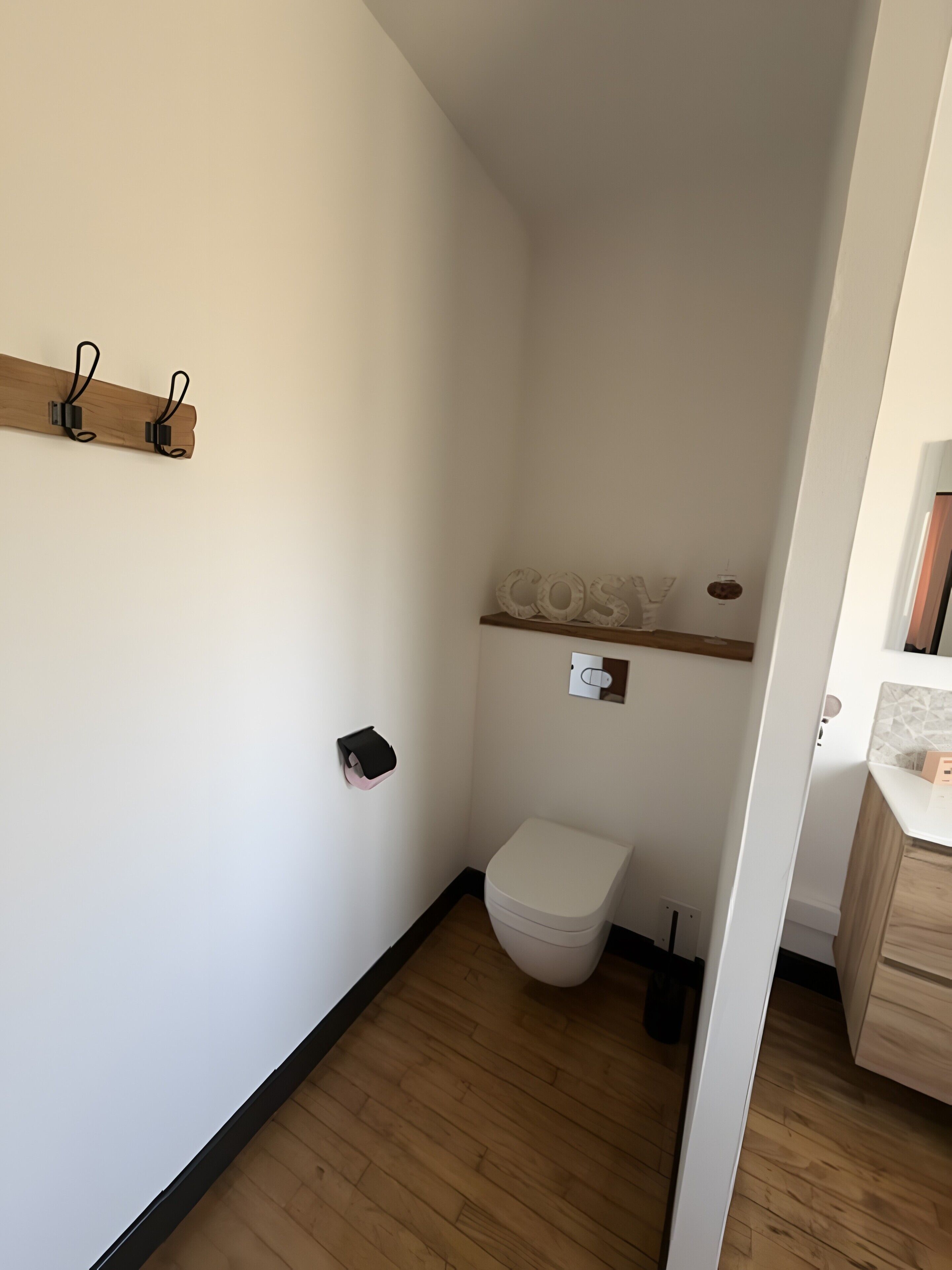 Et lille, smalt badeværelse med toilet, en hylde med et håndklæde og trægulv.