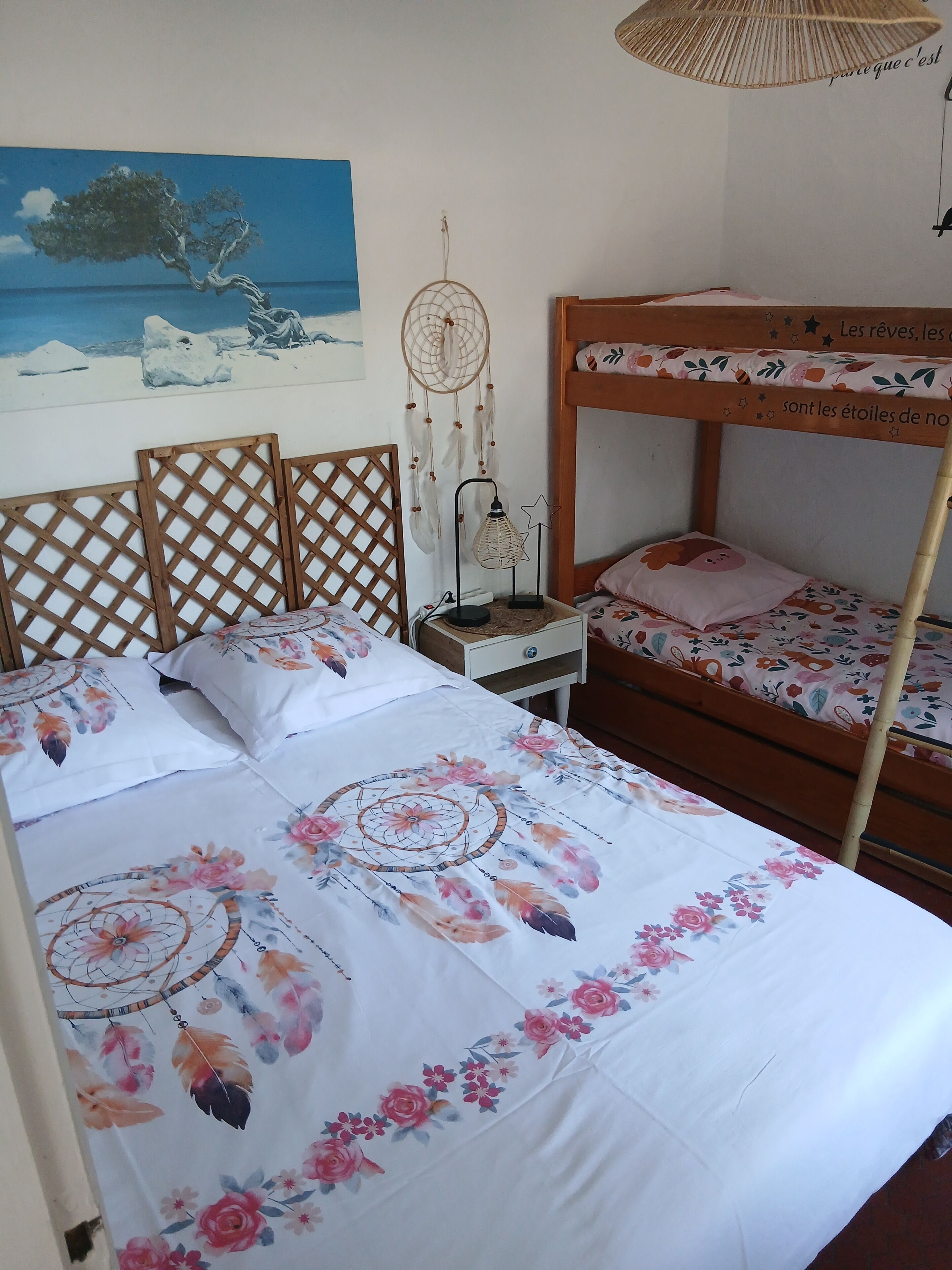 Un dormitorio con una cama litera, una cama individual con colcha estampada de flores, una mesita de noche con un atrapasueños y un cuadro de un árbol en la pared.