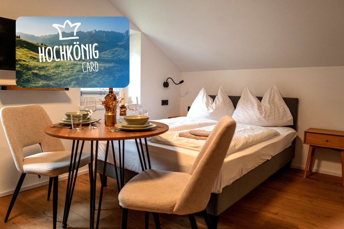 Een slaapkamer met een bed, twee stoelen, een tafel met een vaas en een aan de muur bevestigde televisie waarop 'Hochkönig CARD' staat.