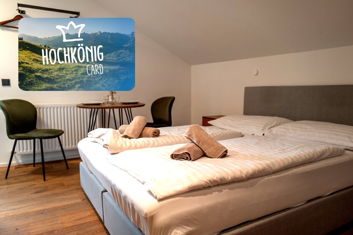 Een hotelkamer met een bed, twee stoelen, een kleine tafel en een wandgemonteerde televisie met 'Hochkönig CARD'.