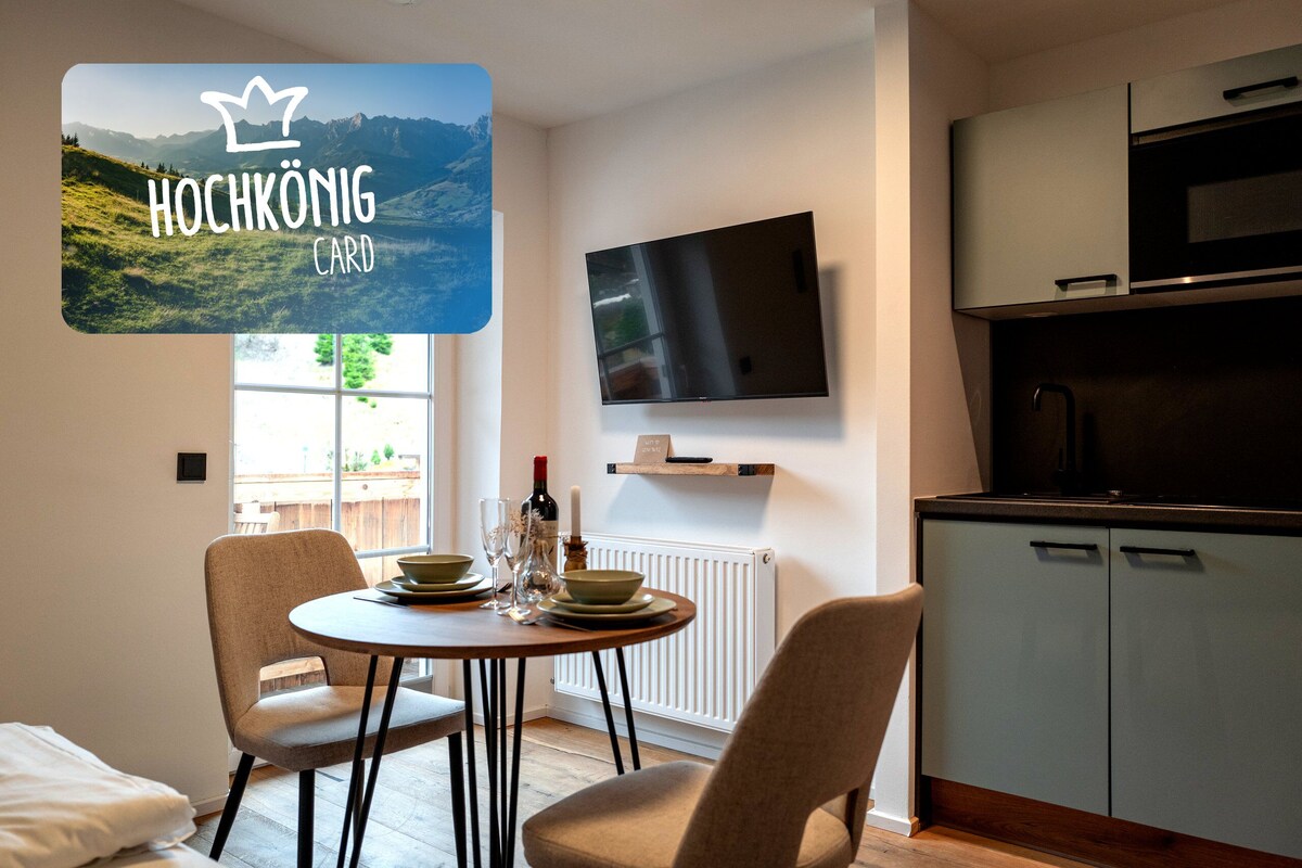 Een moderne kamer met een eettafel, stoelen, een aan de muur bevestigde televisie en een keukenhoek.