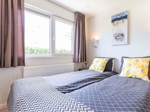 A bedroom with a bed, two pillows, a nightstand, a wall-mounted lamp, and a window with curtains. - Holiday Home in Noordwijk With Terrace (Noordwijk)