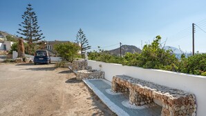 Una carretera costera con un coche estacionado al borde, bancos de piedra a lo largo del camino y una vista despejada del mar y montañas lejanas.