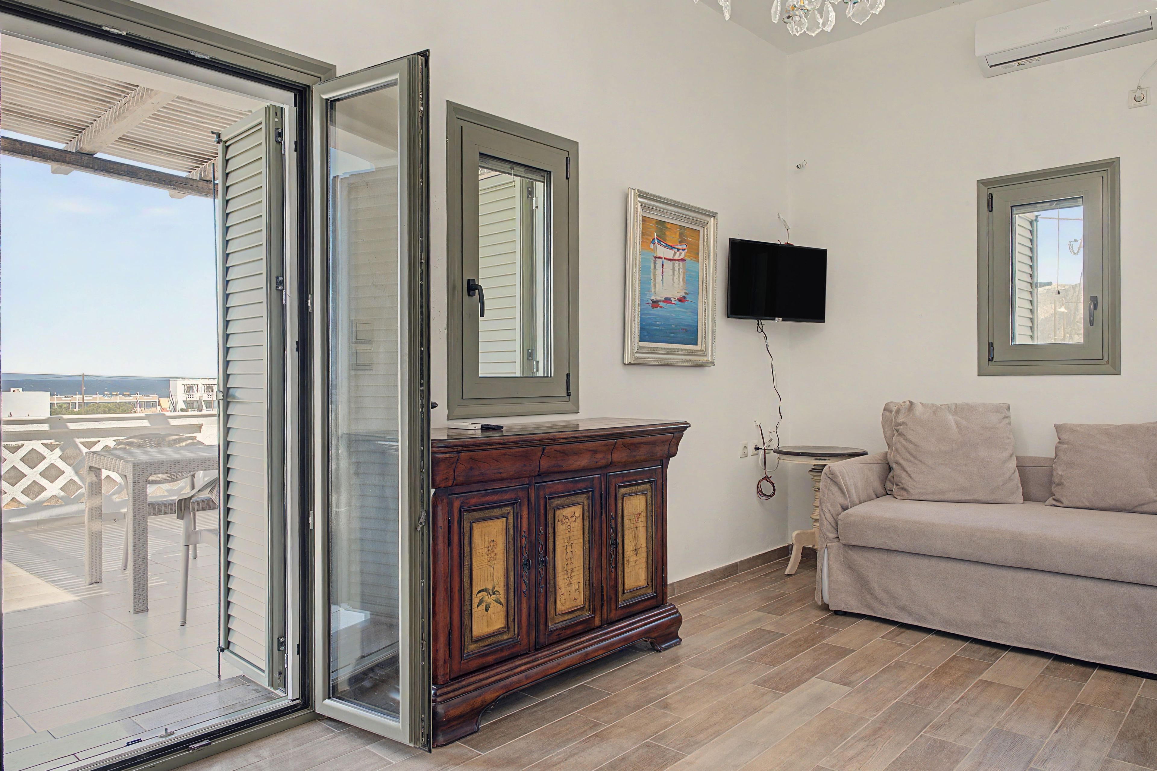 Una stanza con una porta finestra scorrevole che dà su un balcone, un armadio in legno, una TV a schermo piatto fissata al muro e un divano color beige.