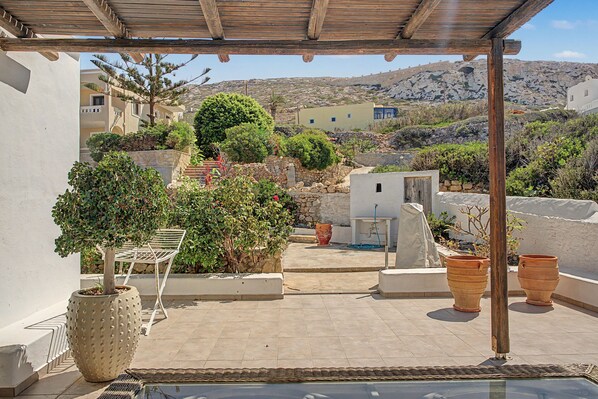 A covered outdoor patio with a view of a hillside, stone walls, and potted plants.