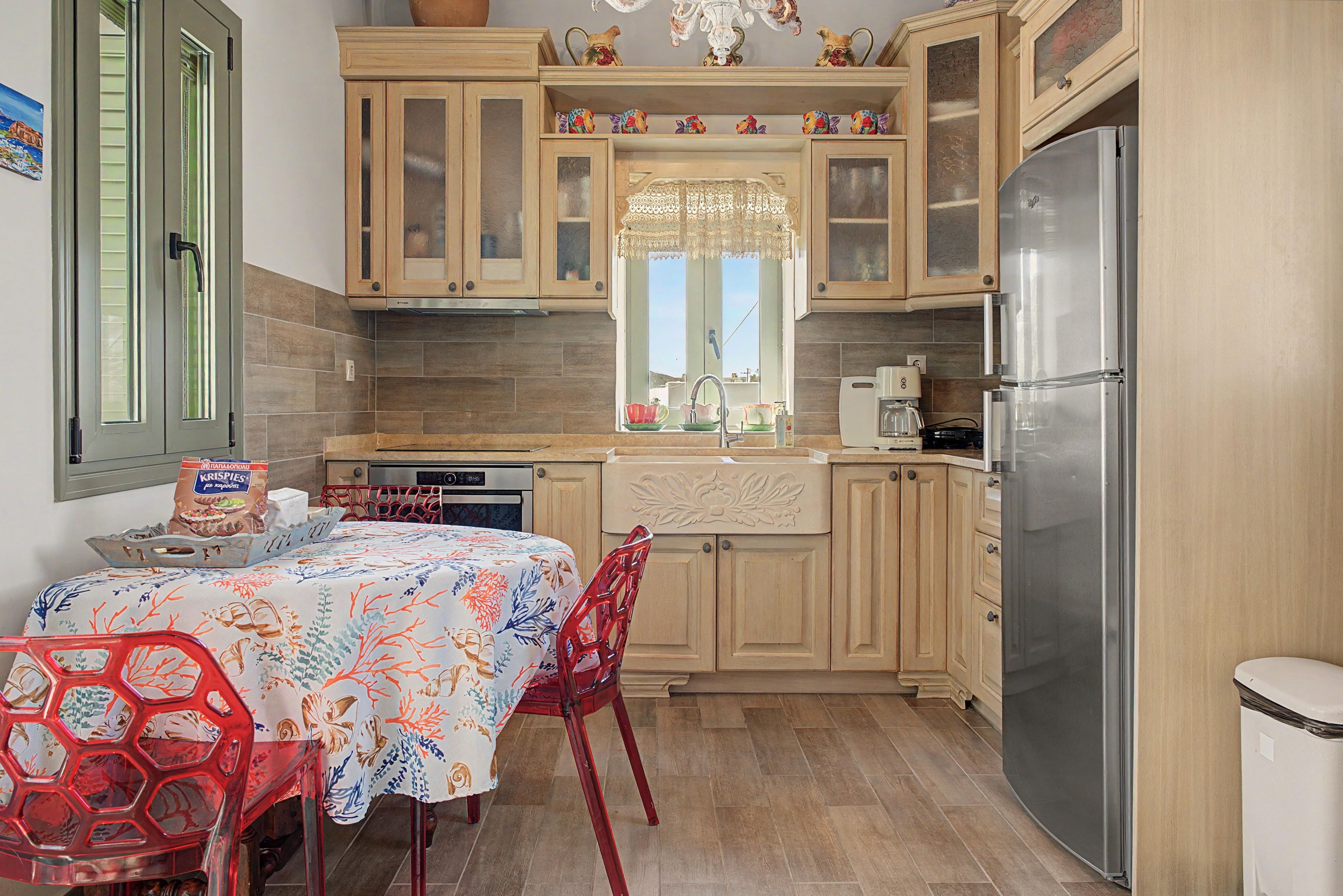 A compact kitchen with wooden cabinets, a stainless steel refrigerator, a dining table with chairs, and a window with a decorative curtain.