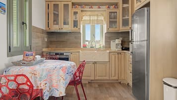 A compact kitchen with wooden cabinets, a stainless steel refrigerator, a dining table with chairs, and a window with a decorative curtain.
