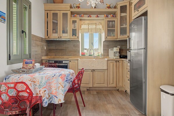 A compact kitchen with wooden cabinets, a stainless steel refrigerator, a dining table with chairs, and a window with a decorative curtain.