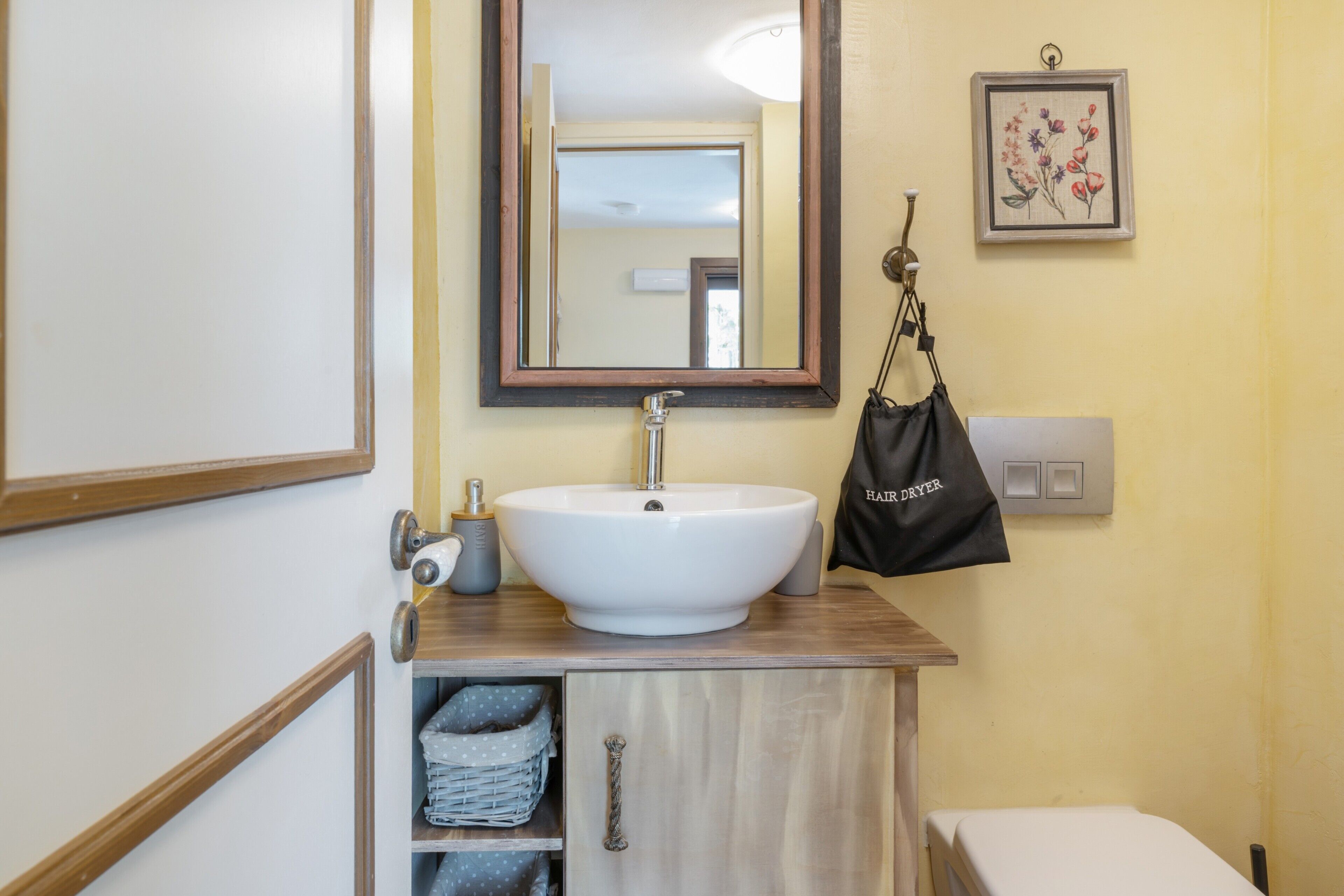 Un baño con lavamanos blanco, tocador de madera, un espejo, un cuadro con flores enmarco y un bolso negro colgado en la pared.