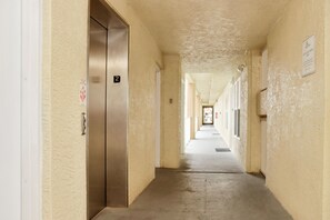 Ein Korridor mit einem metallenen Aufzug, nummerierten Türen und einem Schild an der Wand.