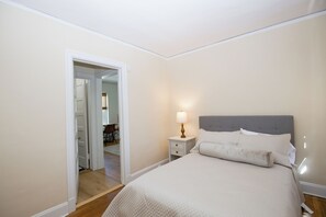 A bedroom with a bed, a nightstand, a lamp, and a door leading to another room. - Broad Street 2 BR Apartment in the Heart of Downtown Southern Pines (Southern Pines)