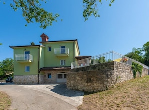 Un bâtiment à deux étages, doté d’un escalier en pierre menant à une porte, d’un balcon au deuxième étage et d’un mur en pierre surmonté d’une rambarde métallique.