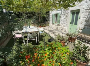 Ein Steinhaus mit grüner Tür und Fensterläden, einem runden Tisch mit Stühlen und einem Garten mit Blumen.