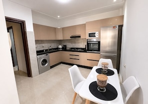 A compact kitchen with a washing machine, microwave, oven, and refrigerator. There is a dining table with two chairs and a decorative bowl. - Modern 2-Bedroom Apartment in Central Gueliz Near Carré Eden (Marrakesh)