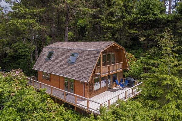 Charming cabin nestled in a lush forest setting, featuring spacious wrap-around deck and classic barn-style architecture surrounded by towering evergreens.