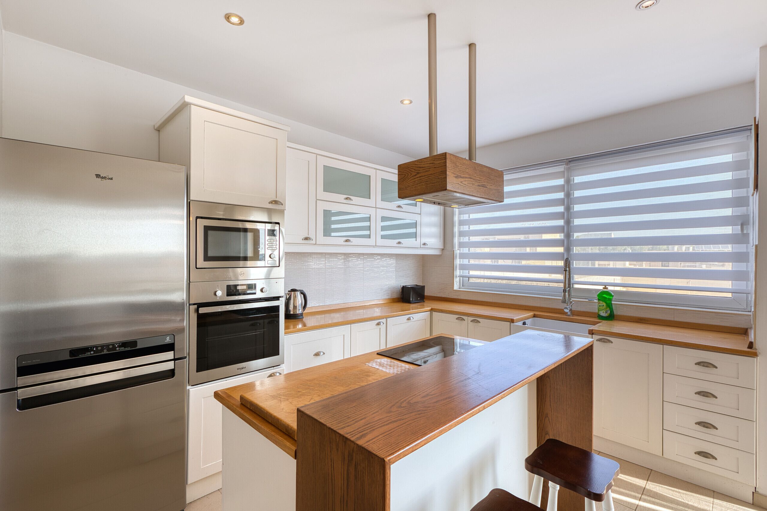 Una cocina moderna con electrodomésticos de acero inoxidable, una isla de madera y un amplio ventanal con persianas.