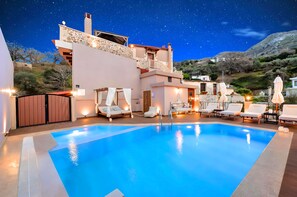 A swimming pool with lounge chairs and umbrellas, surrounded by a building with stone and stucco exterior walls, under a starry night sky. - Petroktisto Luxury Villa Heated Pool & Jaccuzzi (Myriokefala)