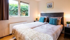 A double bed with a textured duvet, a window with curtains, a bedside table with a lamp, and a framed picture on the wall. - Holiday Home for 5 People (Noordwijk)
