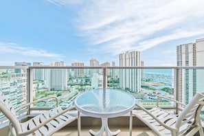 A balcony with a glass table and white chairs, overlooking a cityscape with high-rise buildings. - Newly Renovated Ocean with Queen Bed (Honolulu)