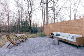 Une terrasse extérieure avec une table et des chaises en bois, un canapé en osier et une poubelle en métal.