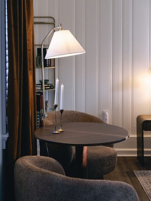 A round table with a lamp, two candles, and a chair in a room with a bookshelf and curtains.