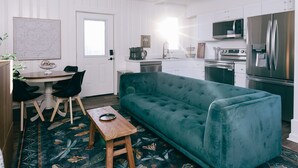 A compact living space with a teal sofa, a dining table with chairs, a kitchen area with modern appliances, and a patterned rug.