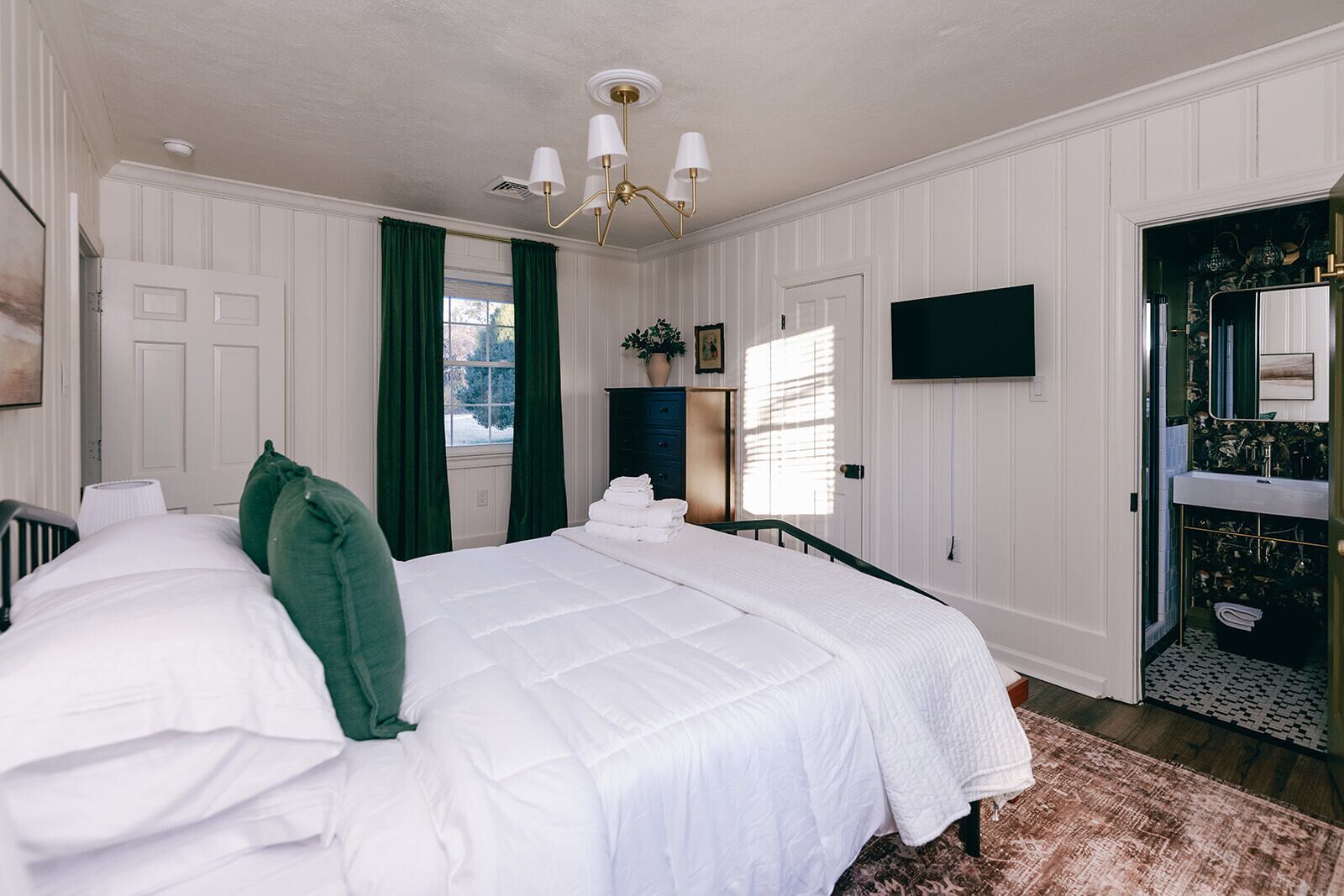 A bedroom with a bed, a dresser, a TV, and a bathroom visible through an open door.