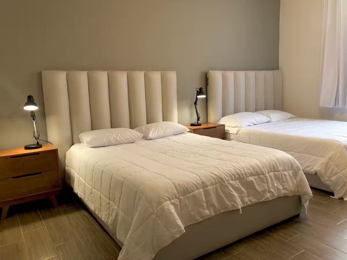 Une chambre d’hôtel avec deux lits, une table de chevet en bois, une lampe et une tête de lit fixée au mur.