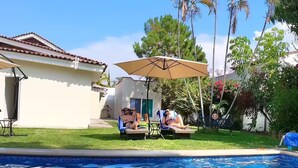 Une piscine avec deux chaises longues sous un auvent, entourée d’une pelouse et de palmiers.