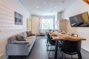 A modern dining area with a wooden table set for four, a grey sofa, and a TV mounted on the wall. - Two-Floor Apartment in Levi Center (Kittilä)