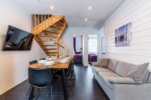 A dining area with a wooden table set for four, a grey sofa, a wooden staircase, and a mounted picture on the wall. - Two-Floor Apartment in Levi Center (Kittilä)